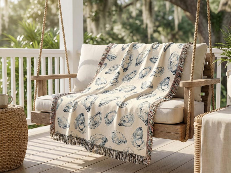 May include: A cream-coloured throw blanket with a blue oyster shell pattern draped over a wooden swing seat. The blanket has a fringed edge and is on a porch with a wicker side table and a mug.