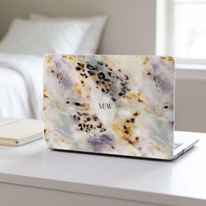 May include: A laptop computer featuring a protective skin with a marbled pattern in shades of cream, white, and soft purple, accented by black leopard print spots. The center of the laptop lid displays the monogram letters M|W in a thin font.