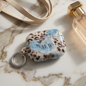 May include: A protective case for wireless earbuds featuring a marbled pattern in shades of blue, cream, and brown with a leopard print design. The case is personalized with the black initials M|W and includes a silver-toned metal ring attachment.