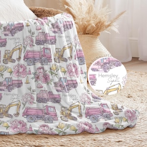 May include: A white throw blanket featuring a repeating pattern of pink dump trucks, yellow excavators, orange traffic cones, and pink floral blossoms. The text Hemsley Sophie is printed in a script font throughout the design of the blanket.