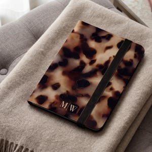 May include: A rectangular notebook featuring a brown and black tortoiseshell pattern on the cover. The notebook includes a black elastic band closure and the letters M/W printed in a gold-toned font near the bottom right corner of the cover.