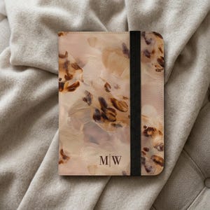 May include: A rectangular notebook with a beige and brown abstract watercolour pattern on the cover. The cover features the initials M|W in a dark brown serif font at the bottom centre and is secured by a vertical black elastic band.
