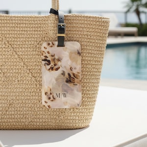 May include: A beige woven straw tote bag featuring a rectangular luggage tag made of marbled acrylic in shades of cream, tan, and dark brown. The tag displays the letters MW in a black serif font and attaches with a black leather strap and buckle.
