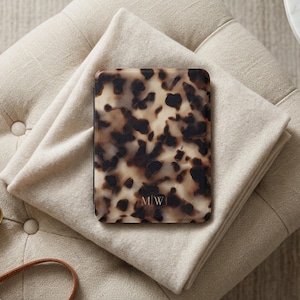 May include: A rectangular e-reader case featuring a brown, black, and cream tortoiseshell pattern. The bottom centre of the case displays the letters M|W in a small, thin, white serif font. The case has rounded corners and a smooth, glossy finish.