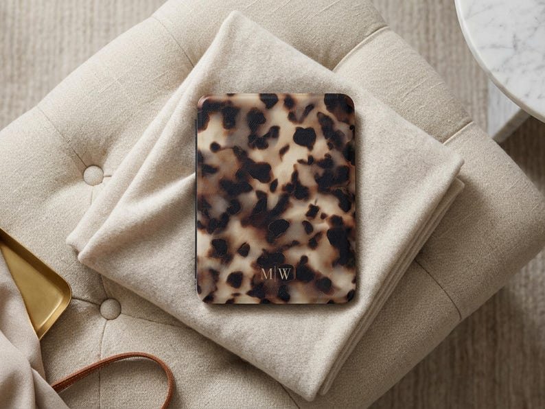 May include: A rectangular e-reader case featuring a brown, black, and cream tortoiseshell pattern. The bottom centre of the case displays the letters M|W in a small, thin, white serif font. The case has rounded corners and a smooth, glossy finish.