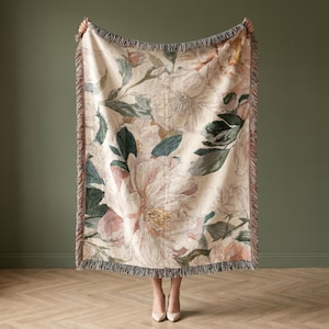 May include: A floral throw blanket with a cream background and large pink and white flowers. The blanket has green leaves and a fringed edge. The blanket is held up against a dark green wall and light wood floor.