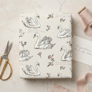 Flat lay of personalized swan wrapping paper with gold scissors blush silk ribbon and wedding gift card on a stone surface showing elegant bridal shower gift wrap design