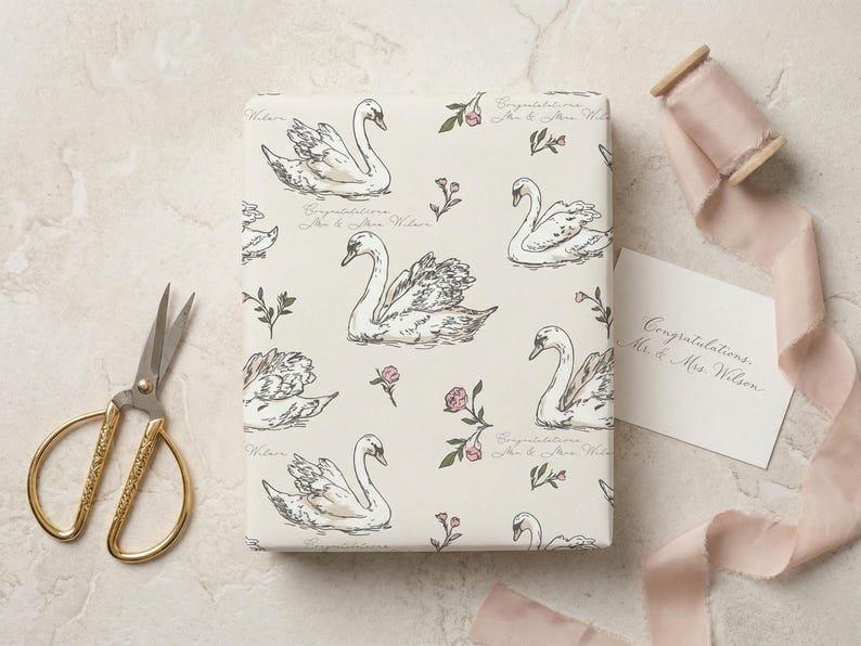 Flat lay of personalized swan wrapping paper with gold scissors blush silk ribbon and wedding gift card on a stone surface showing elegant bridal shower gift wrap design