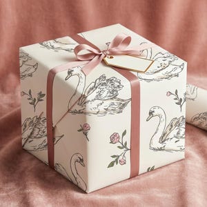 Gift box wrapped in romantic swan wedding paper with blush satin ribbon and blank tag styled on pink velvet for feminine bridal shower gift wrap presentation