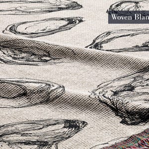 May include: A woven blanket with a cream-colored base featuring a repeating black line-art pattern of oyster shells. The fabric has a textured, waffle-like weave and includes the text Woven Blanket in the upper right corner with colorful fringe.