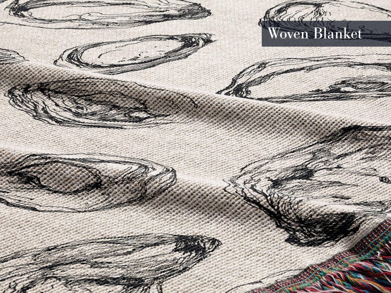 May include: A woven blanket with a cream-colored base featuring a repeating black line-art pattern of oyster shells. The fabric has a textured, waffle-like weave and includes the text Woven Blanket in the upper right corner with colorful fringe.