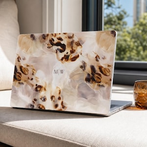 May include: A laptop computer featuring a protective cover with an abstract pattern of swirling cream, beige, and dark brown tones. The center of the cover displays the monogram M|W in a simple, thin, dark-colored serif font against the light background.