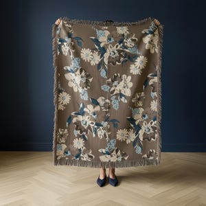 May include: A brown floral throw blanket with a fringe edge. The blanket features large white, blue, and brown flowers. The throw is held up against a dark blue wall.