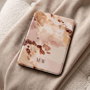 May include: A rectangular e-reader cover featuring an abstract watercolor pattern in shades of beige, cream, and brown. The design includes dark brown ink-like splatters and the letters M/W printed in a simple serif font at the bottom center.