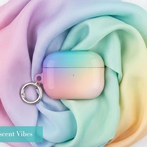 May include: A protective case for wireless earbuds featuring a smooth, gradient finish that transitions from soft pink to yellow, green, and blue. The case includes a silver-toned metal ring attachment on the side and displays the text Iridescent Vibes.
