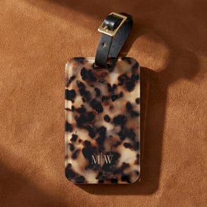May include: A rectangular luggage tag featuring a brown and black tortoiseshell pattern with the letters MW printed in gold at the bottom. The tag includes a black leather strap with a gold-toned metal buckle for secure attachment to luggage.