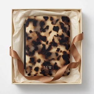 May include: A rectangular e-reader case featuring a brown and black tortoiseshell pattern. The bottom centre of the case displays the letters M|W in a gold-toned font. The case is presented inside a square gift box with cream tissue paper.