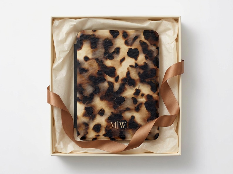 May include: A rectangular e-reader case featuring a brown and black tortoiseshell pattern. The bottom centre of the case displays the letters M|W in a gold-toned font. The case is presented inside a square gift box with cream tissue paper.