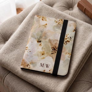 May include: A rectangular notebook with a cream-colored cover featuring an abstract watercolor pattern in shades of beige, soft pink, and sage green. The cover displays the letters M/W in dark brown and includes a vertical black elastic band.
