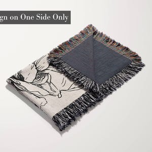 May include: A woven throw blanket featuring a black line art floral illustration on a light beige background. The blanket has a dark gray reverse side and multicolored fringe edges. The text "Design on One Side Only" is visible.