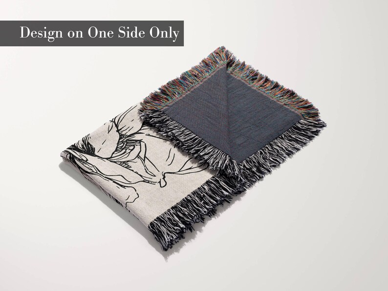 May include: A woven throw blanket featuring a black line art floral illustration on a light beige background. The blanket has a dark gray reverse side and multicolored fringe edges. The text "Design on One Side Only" is visible.