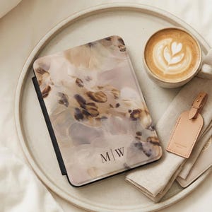 May include: A journal with a floral design and the initials "MW" is displayed on a round tray with a cup of coffee, a luggage tag, and a passport holder. The journal cover has a beige, brown, and white pattern.