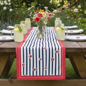 Puede incluir: Una mesa de picnic de madera preparada para una reunión de verano. La mesa está adornada con un camino de mesa a rayas rojas, blancas y azules con detalles de estrellas. Frascos de vidrio transparentes llenos de limonada y flores frescas.