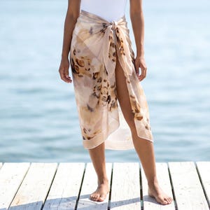 May include: A sheer, lightweight sarong wrap featuring an abstract, watercolor-style print in shades of beige, brown, and cream. The fabric is tied at the waist with a simple knot, creating a draped, flowing silhouette that falls to the mid-calf.
