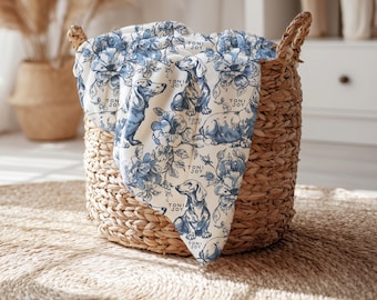 May include: A white fabric blanket featuring a blue floral pattern and illustrations of dachshund dogs. The text TONI JOY is printed repeatedly across the surface. The blanket is draped over a woven brown basket with two handles.
