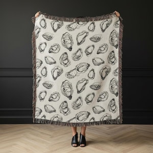 May include: A cream-colored woven throw blanket featuring a repeating black line-art pattern of oyster shells. The rectangular blanket is finished with a dark gray fringed edge along all four sides, creating a textured border around the print.