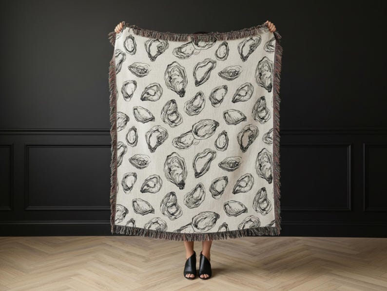 May include: A cream-colored woven throw blanket featuring a repeating black line-art pattern of oyster shells. The rectangular blanket is finished with a dark gray fringed edge along all four sides, creating a textured border around the print.