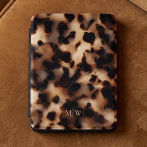 May include: A rectangular e-reader case featuring a brown, black, and cream tortoiseshell pattern. The bottom center of the case displays the gold-colored monogram letters M and W separated by a vertical line, all enclosed in a thin frame.