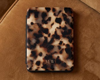 May include: A rectangular e-reader case featuring a brown, black, and cream tortoiseshell pattern. The bottom center of the case displays the gold-colored monogram letters M and W separated by a vertical line, all enclosed in a thin frame.