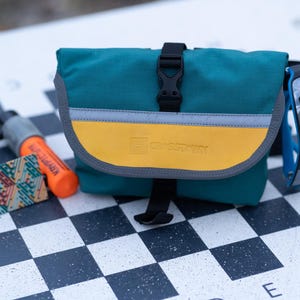 Könnte beinhalten: Türkis-gelbe Umhängetasche mit schwarzem Schnallenverschluss und reflektierendem Streifen. Auf der Tasche steht der Markenname "SWISSWIN". Sonnenbrille und ein orangefarbenes Werkzeug liegen neben der Tasche.