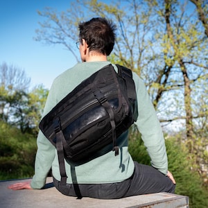 Puede incluir: Un bolso mensajero negro con correas ajustables se lleva sobre el hombro. El bolso está hecho de un material duradero y tiene múltiples compartimentos. La persona lleva una camisa verde de manga larga y pantalones oscuros.