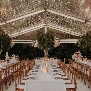 May include: A long, white tablecloth covers a table set for a wedding reception. The table is surrounded by wooden chairs with white cushions. The ceiling is decorated with fairy lights.