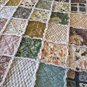 May include: A patchwork quilt featuring a variety of square fabric blocks with diverse floral, geometric, and solid patterns in shades of green, blue, yellow, and purple. Each square is joined with exposed, frayed white seams in a rag quilt style.