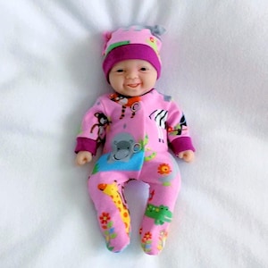 May include: A baby doll wearing a pink onesie and matching hat. The onesie features animal prints including a giraffe, zebra, and hippo. The doll is lying on a white surface and has a smiling expression.