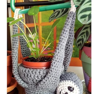 May include: A gray crocheted sloth plant hanger with a white face and a small potted plant hanging from it.