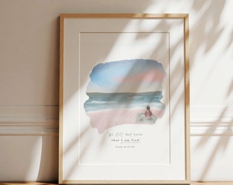 May include: A watercolour painting of a beach scene with a person sitting on the sand. The sky is blue and pink, and the ocean is a light blue. The text "Be still and know that I am God" is written below the painting, along with the reference "Psalm 46:10 NIV".