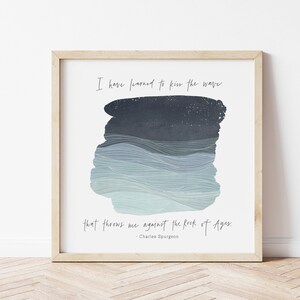 Op de afbeelding: Een ingelijst print met een blauw en wit aquarelontwerp van golven. De quote "Ik heb geleerd om de golf te kussen die me tegen de Rots der Eeuwen werpt. - Charles Spurgeon" staat onder het ontwerp.
