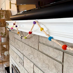 May include: A colorful garland with felt balls in various colors, strung on a natural wooden cord. The garland is draped across a white mantel above a fireplace. The felt balls include red, orange, yellow, green, blue, purple, and black.