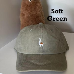 May include: Soft green baseball cap with a small embroidered llama design. The cap has a curved brim and a classic six-panel construction. A brown stuffed animal is in the background. The text "Soft Green" is also in the image.