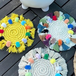 May include: Three colorful crocheted coasters with pom-poms and fringe. The coasters are yellow, white, and green with multi-colored pom-poms.
