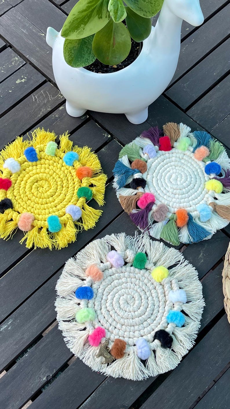 May include: Three colorful crocheted coasters with pom-poms and fringe. The coasters are yellow, white, and green with multi-colored pom-poms.