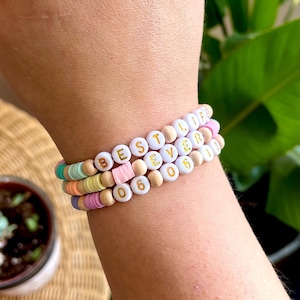 May include: Three stacked bracelets made of colorful polymer clay disc beads and round wooden beads. The bracelets feature white letter beads spelling out "BEST LIFE EVER" and white number beads displaying the date "06 06 22" in gold lettering.