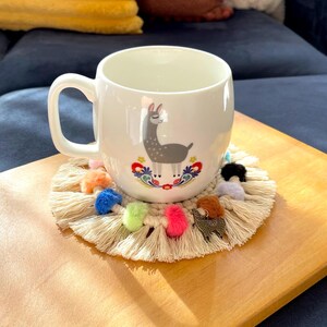 May include: A white ceramic mug with a llama illustration and colorful floral accents sits on a round, beige crocheted coaster with colorful pom-poms. The coaster is on a wooden tray.