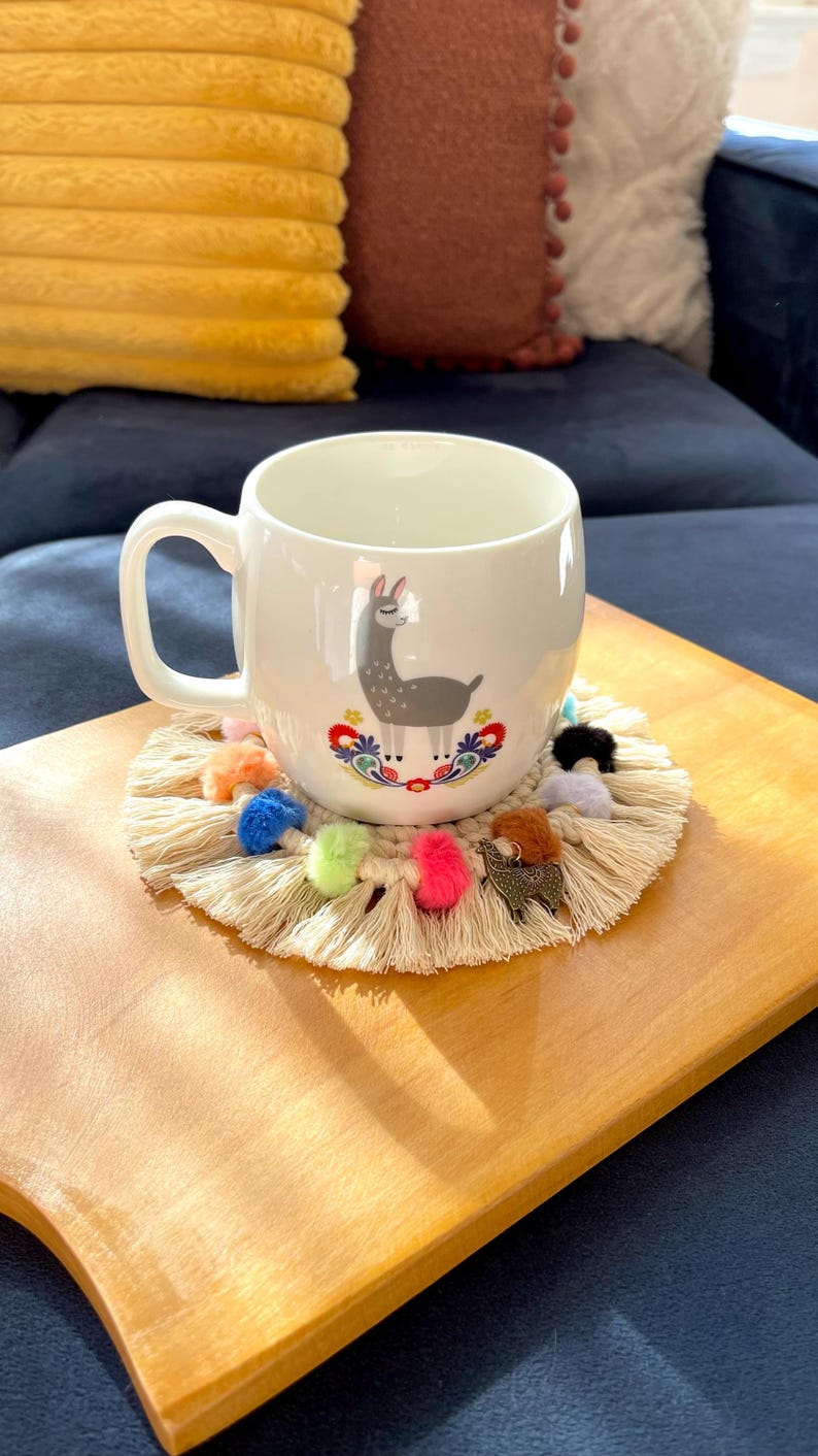 May include: A white ceramic mug with a llama illustration and colorful floral accents sits on a round, beige crocheted coaster with colorful pom-poms. The coaster is on a wooden tray.