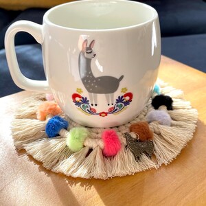 May include: White ceramic mug with a llama illustration and colorful floral accents. The mug is sitting on a beige crocheted coaster with colorful pom-poms and a metal llama charm.
