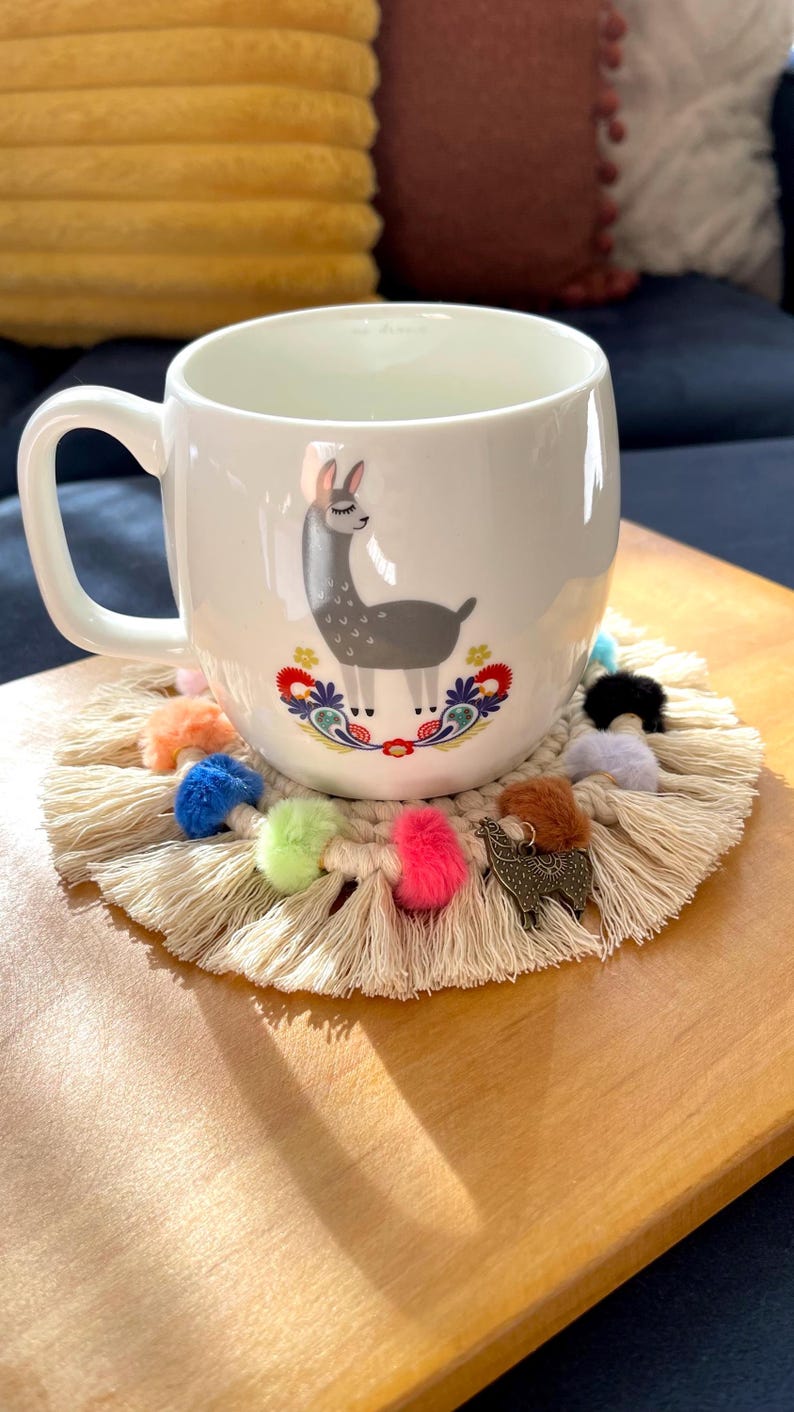 May include: White ceramic mug with a llama illustration and colorful floral accents. The mug is sitting on a beige crocheted coaster with colorful pom-poms and a metal llama charm.